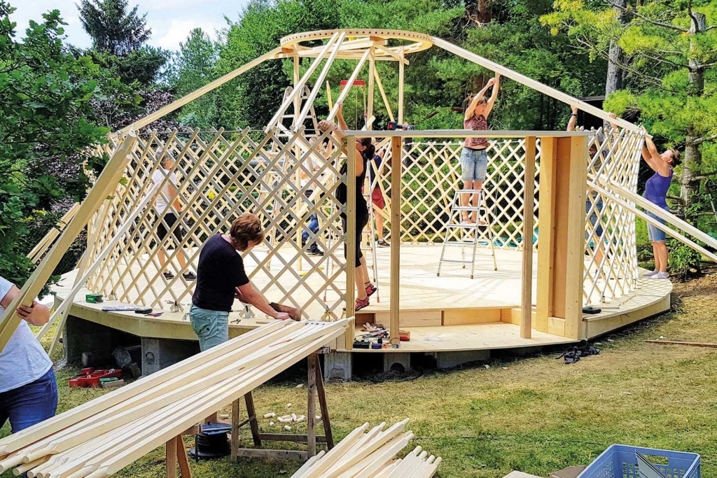 Mehrere Personen bauen eine große, mehrstöckige Holzkonstruktion im Freien mit vielen Holzbalken.