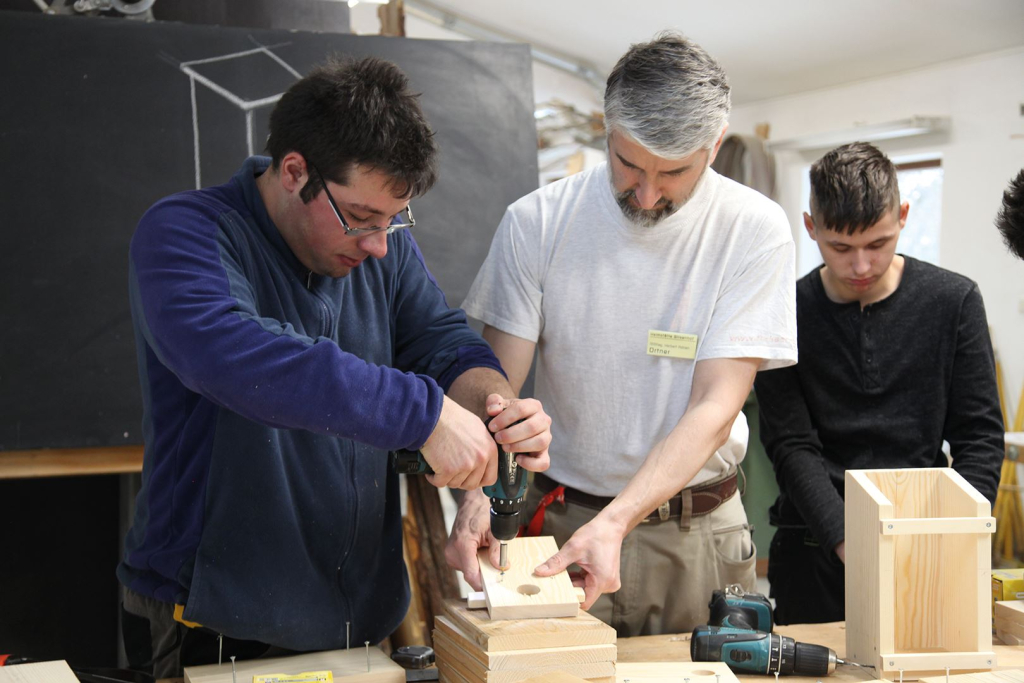 Drei Männer arbeiten in einer Werkstatt an verschiedenen Holzobjekten mit elektrischen Werkzeugen.